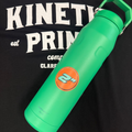 An orange 2 inch round sticker on a green watter bottle laying on t-shirt that says Kinetic Print Co