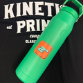 An orange 2 inch square sticker on a green watter bottle laying on t-shirt that says Kinetic Print Co