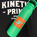 An orange 3 inch square sticker on a green watter bottle laying on t-shirt that says Kinetic Print Co