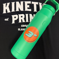 An orange 3 inch round sticker on a green watter bottle laying on t-shirt that says Kinetic Print Co