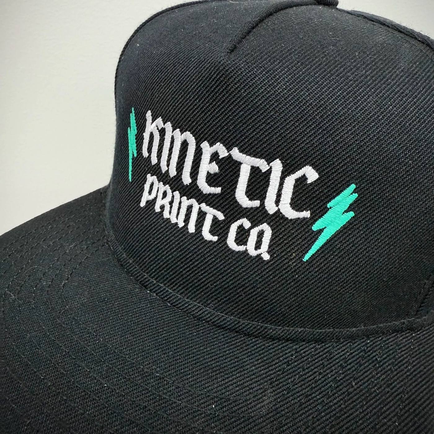 Black trucker hat with embroidered Kinetic Print Co logo and teal lightning bolt